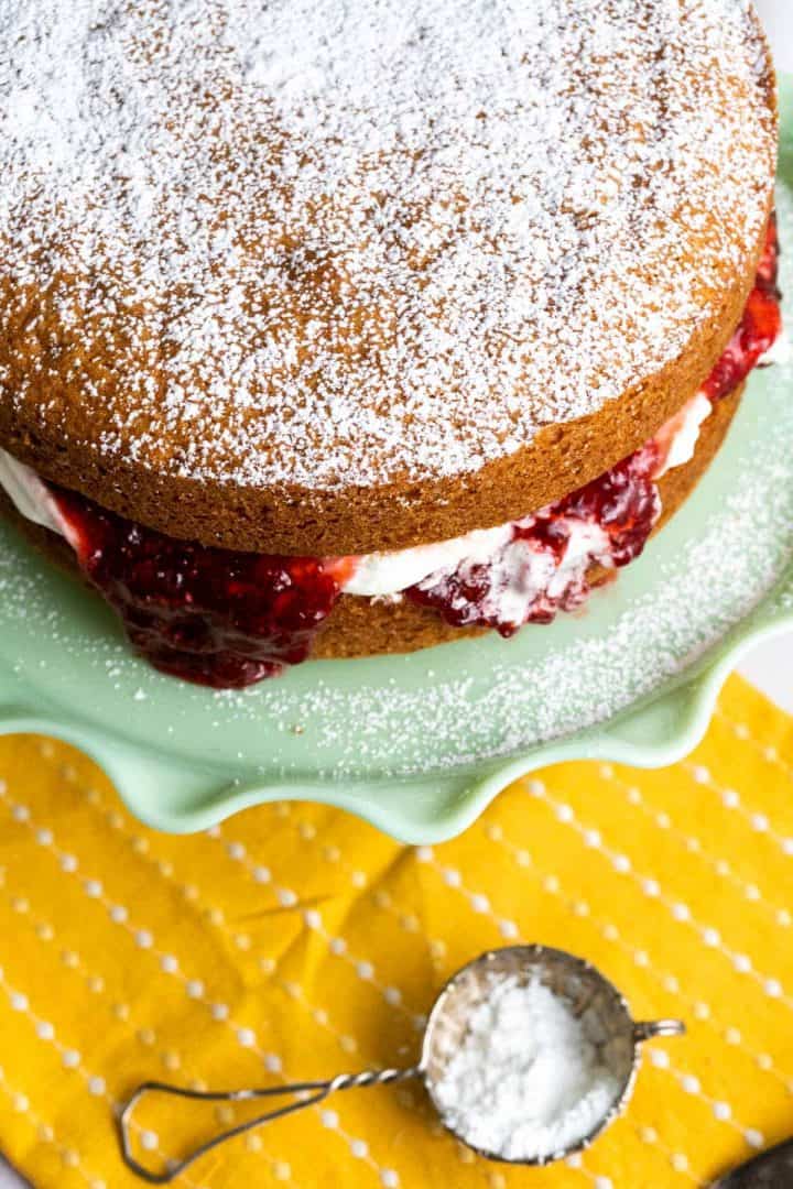 Over the head shot of Victoria Sponge cake with a strainer on the side with powdered sugar in it.