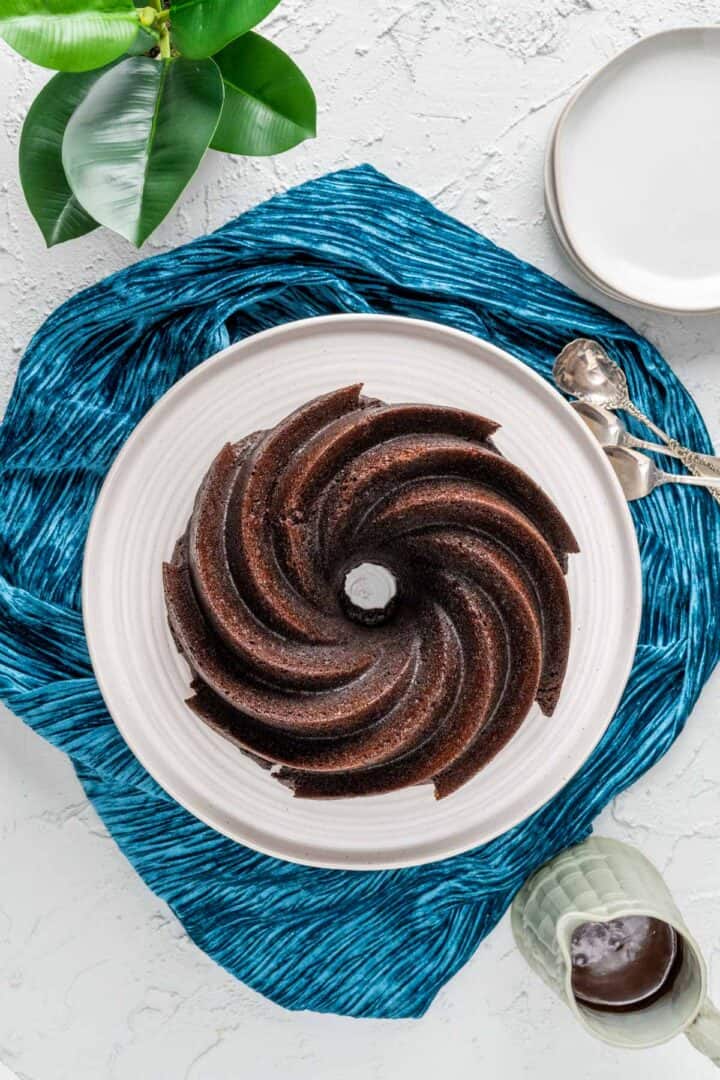 Chocolate bundt cake in the white cake plate with ganache on the side.