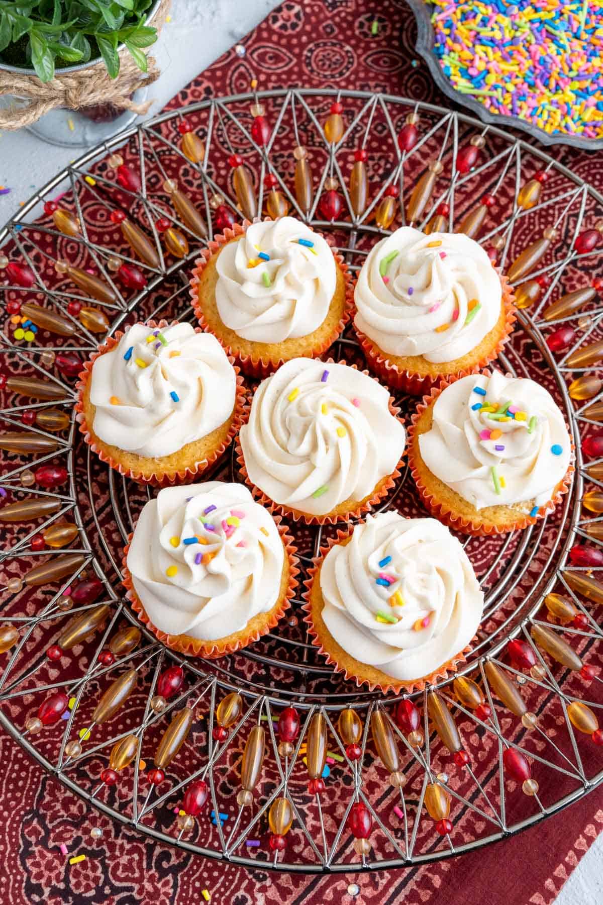 seven vanilla cupcakes with oil on a colorful plate, frosted with butter cream and sprinkles on the top.