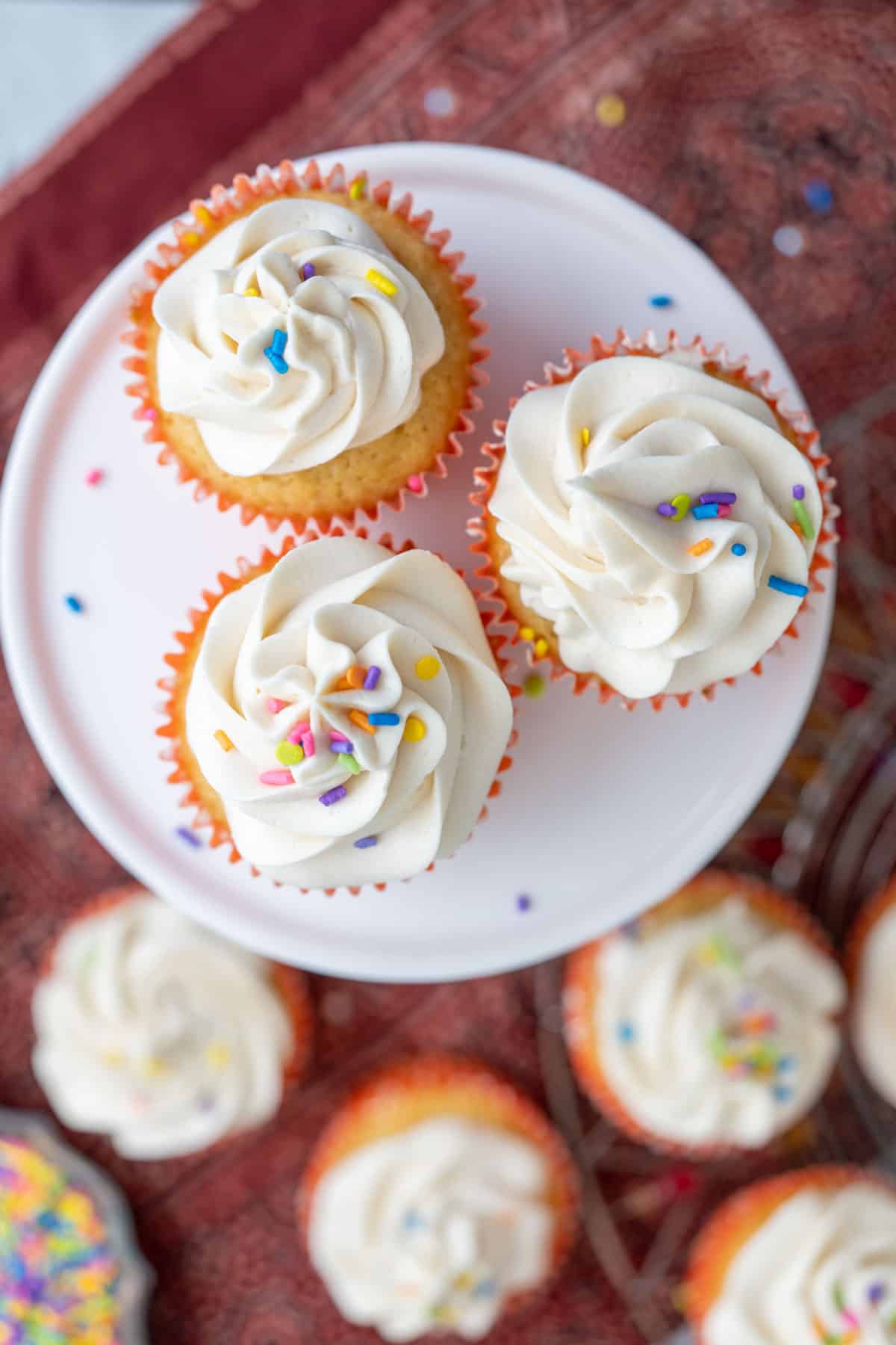 three vanilla cupcakes with oil on a white serving plate on a red table cloth.