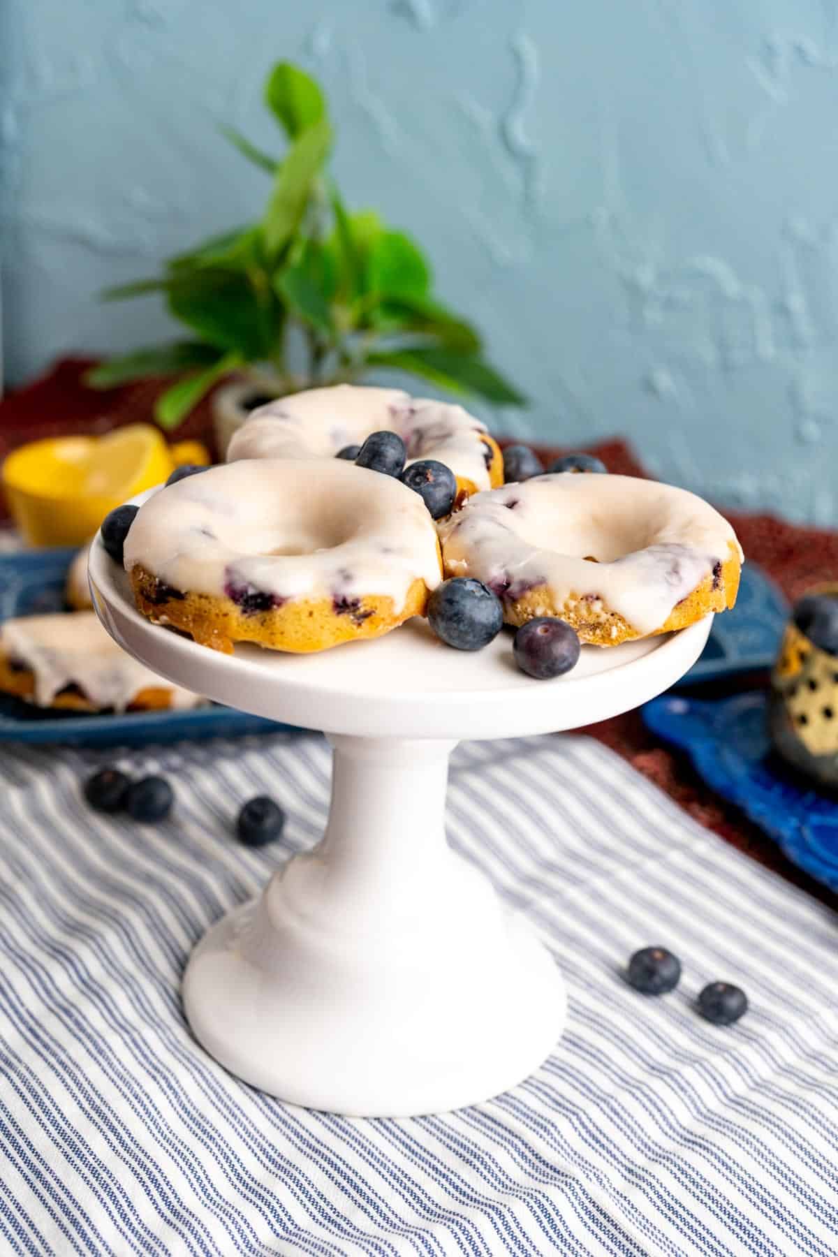 three blueberry donuts on a white stand over a blue table cloth.