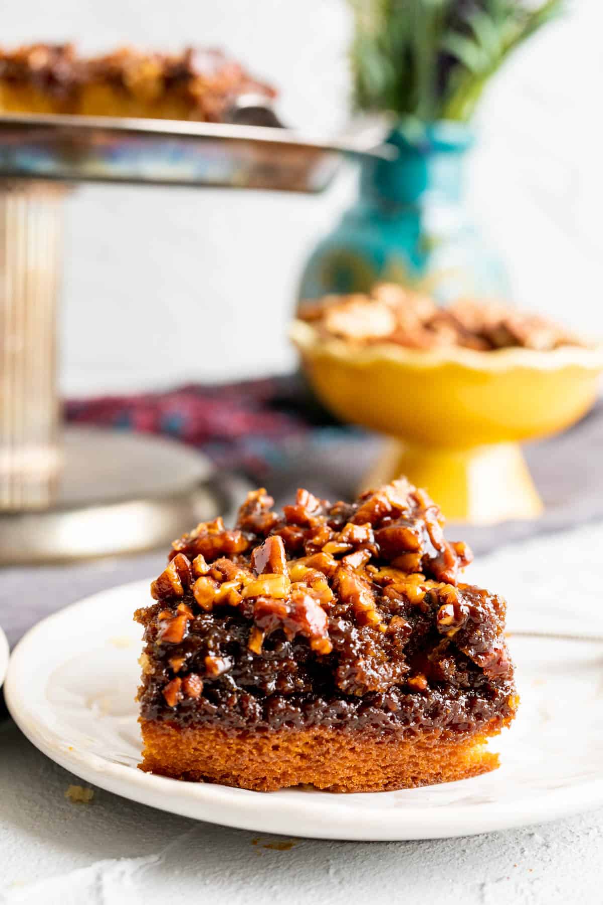 One slice of pecan cake on a white plate with pecans in a stand in the backdrop.