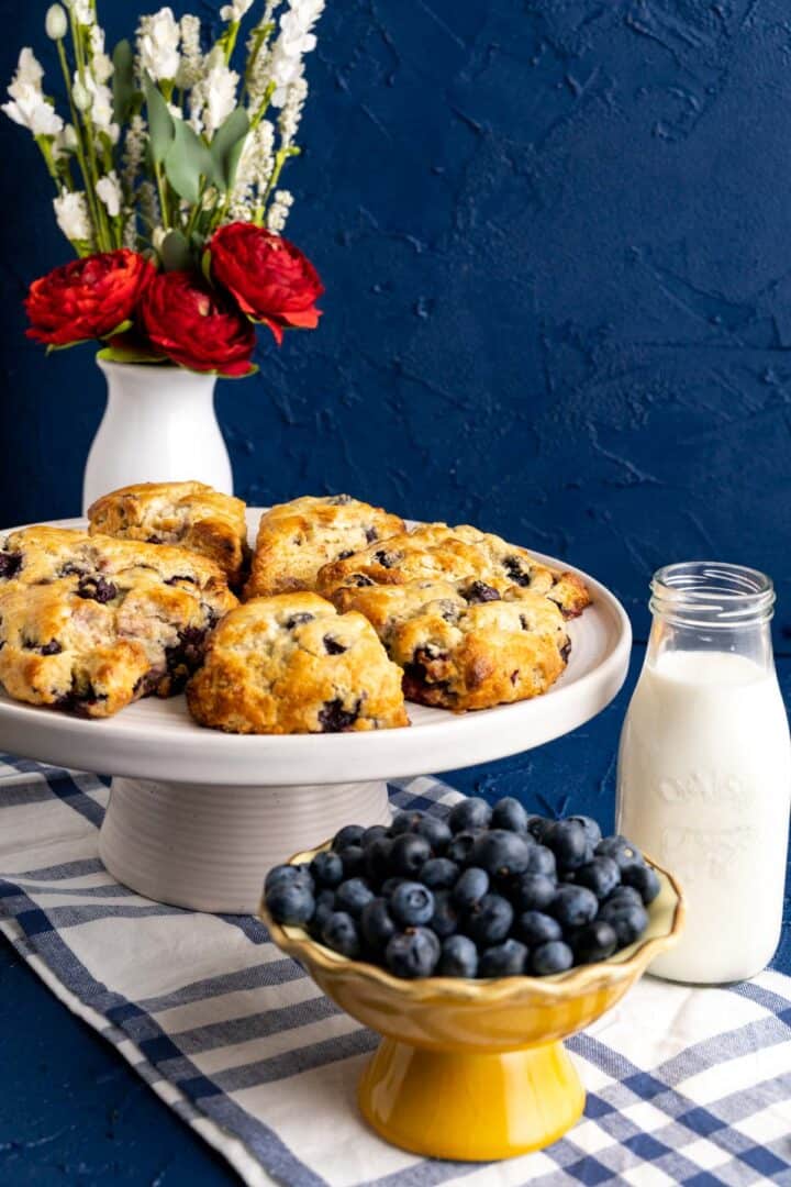 blueberry scones on the big white cake plate with a glass of milk and a bowl of blueberries around.