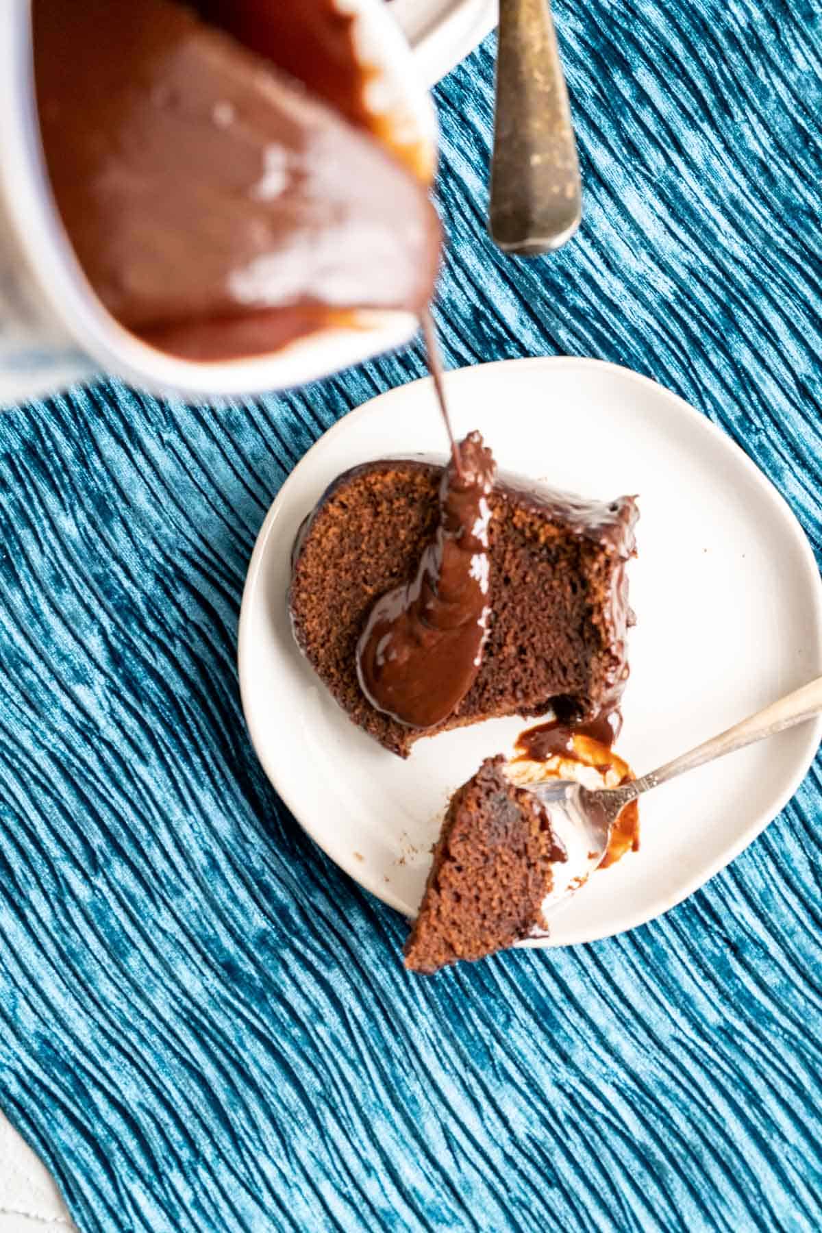 pouring the ganache on a chocolate cake slice sitting in a white plate.