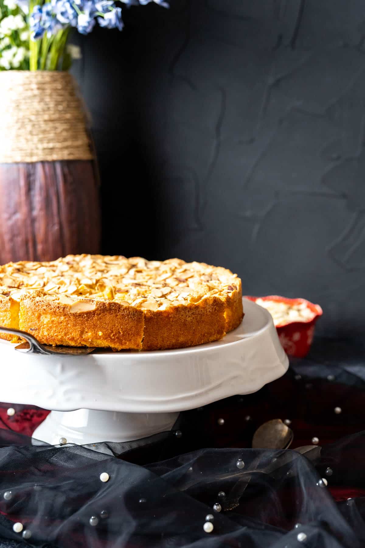 Almond Cake on a white cake plate with a flower vase in the backdrop.