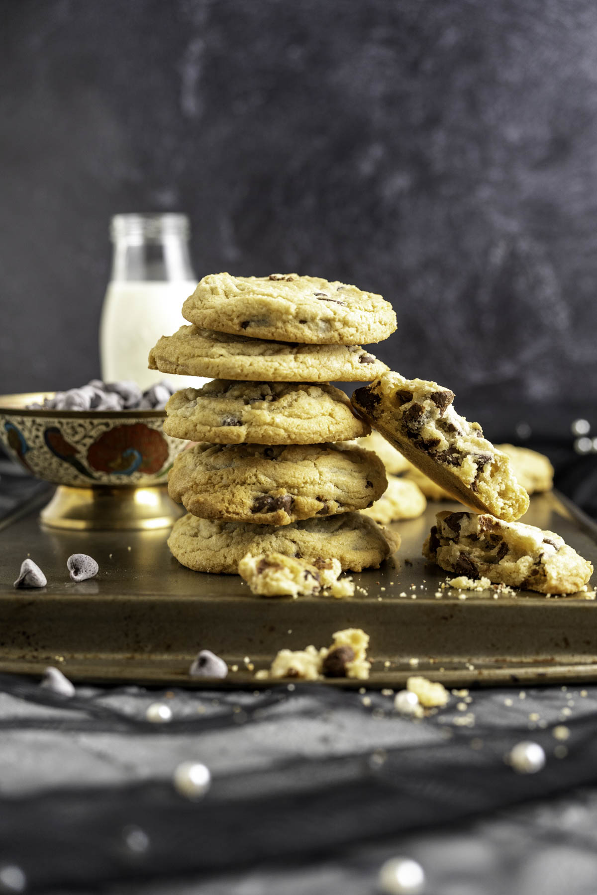 five chocolate chip cookies placed on one another with a bowl of chocolate chips and a milk bottle in the backdrop.