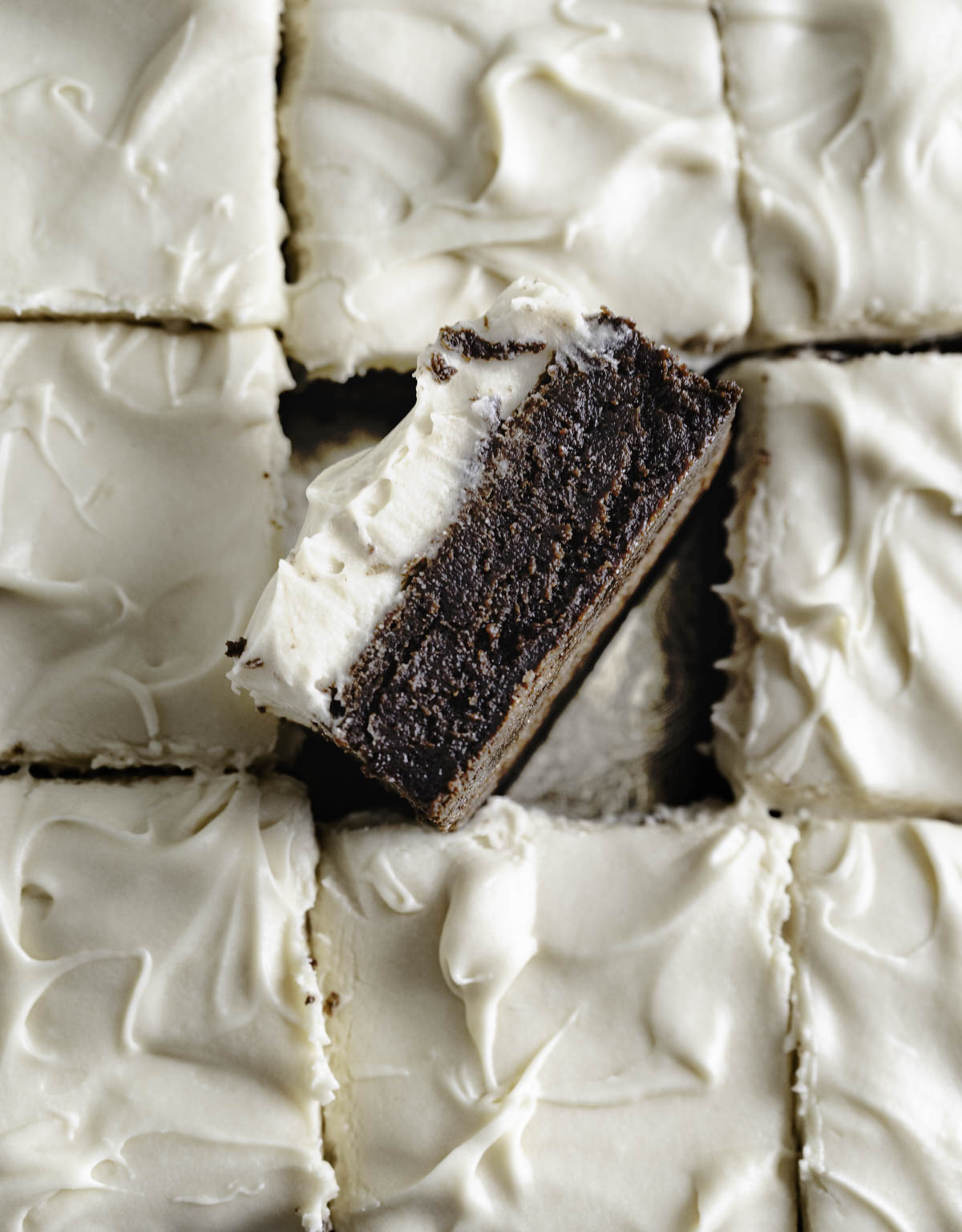 A piece of chocolate brownie with cream cheese frosting standing diagonally among the rest of the pieces.