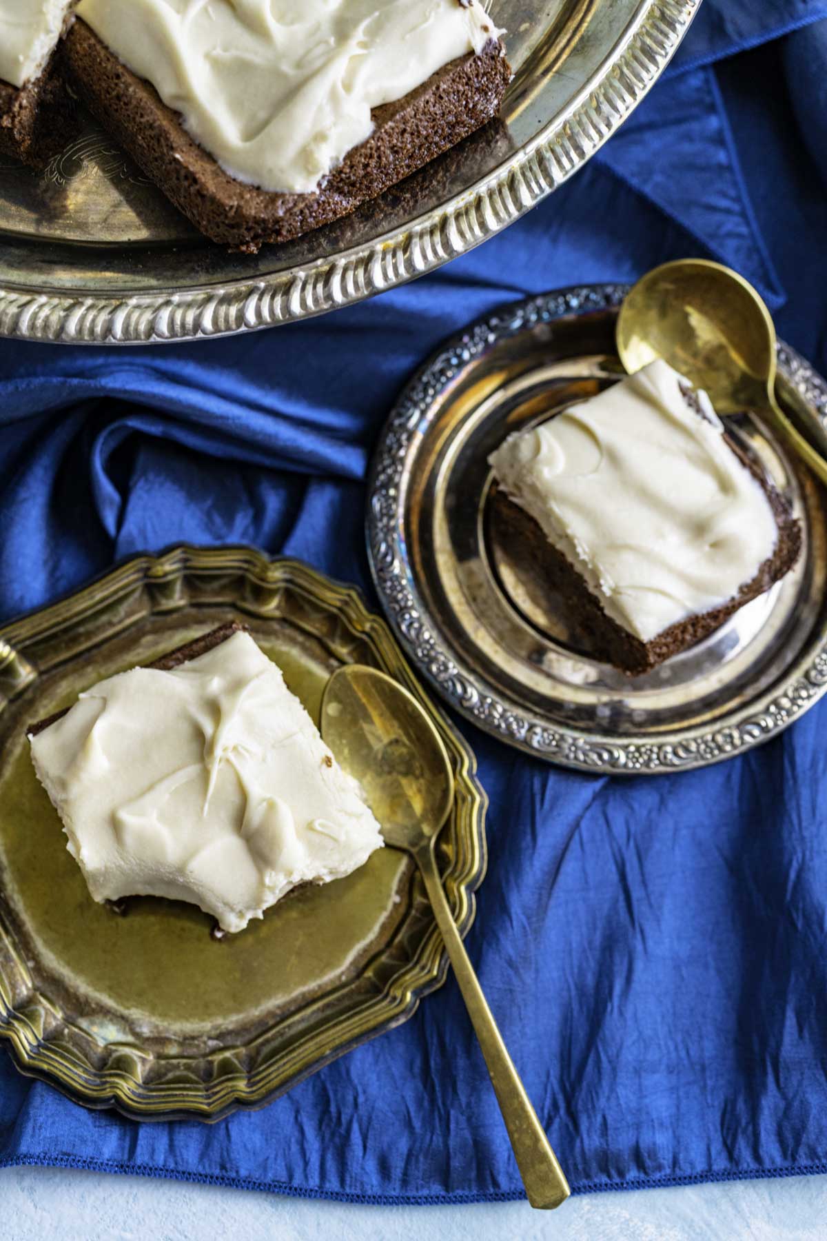 Two brownie pieces with frosting on golden plates on a dark blue table cloth.