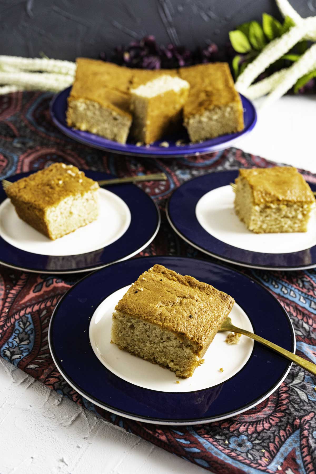 Three Desi Ghee cake pieces plates sitting on gray printed table cloth with golden forks.