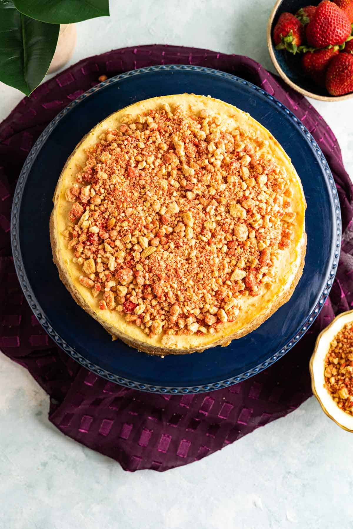 over the head shot of strawberry crunch cheesecake sitting in a dark blue cake plate with strawberries and strawberry crunch in a bowl around.