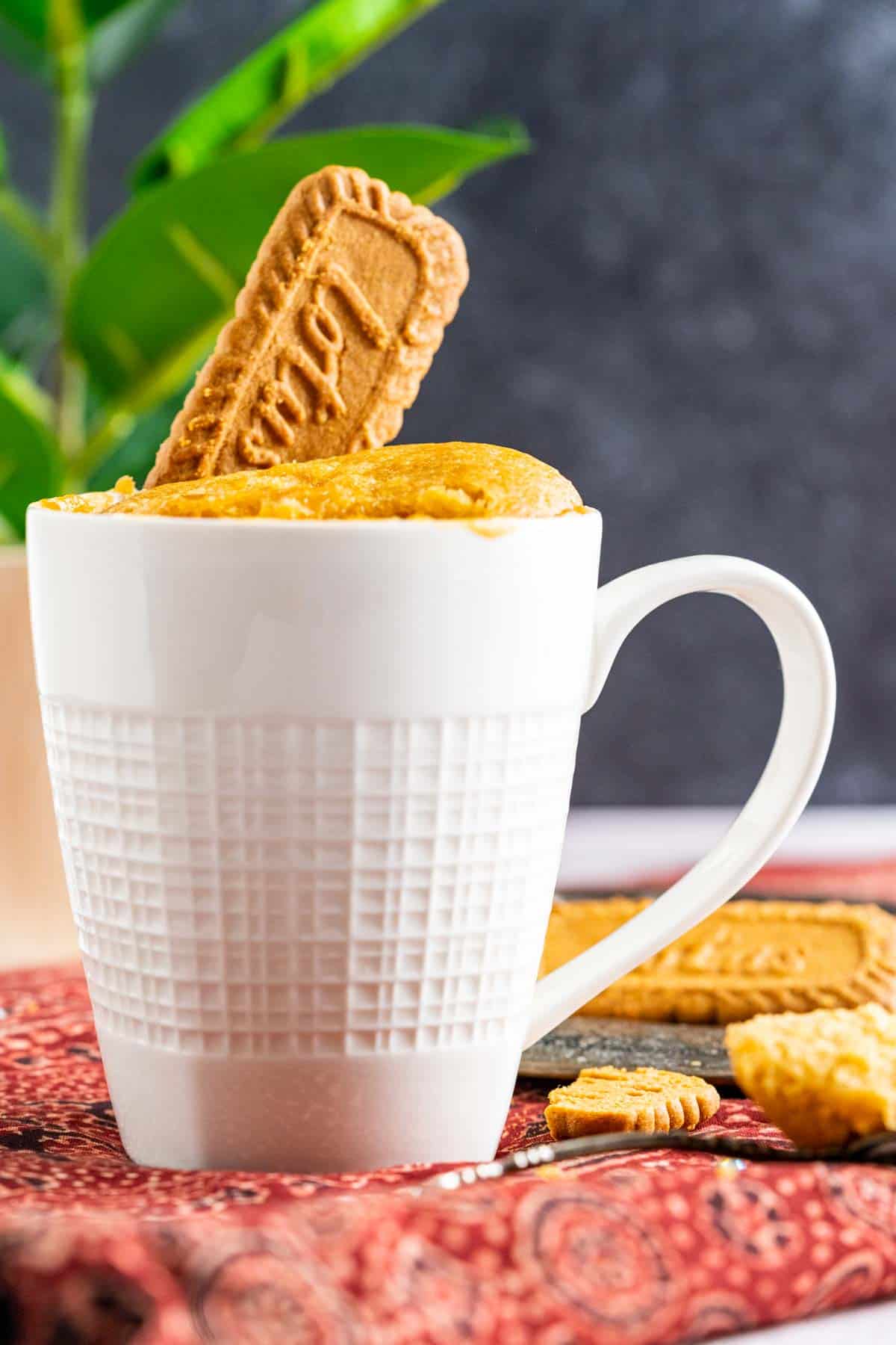 Lotus mug cake in a white mug with lotus cookies spread around on red table cloth.