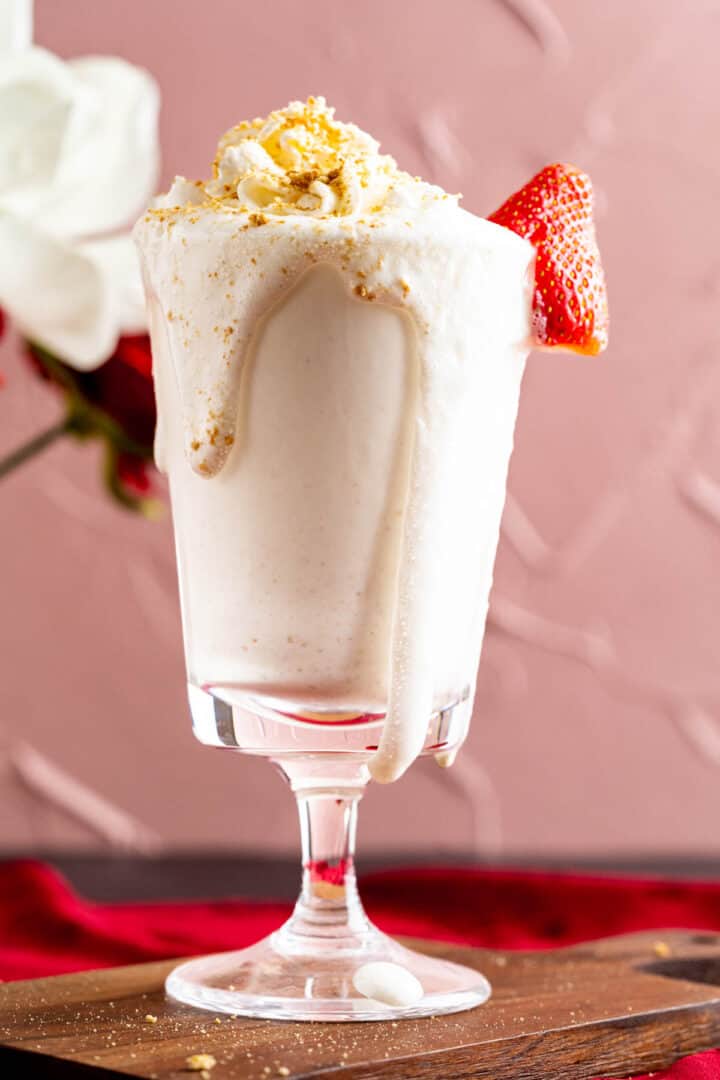 milkshake glass decorated with a strawberry piece sitting over a wooden plate.