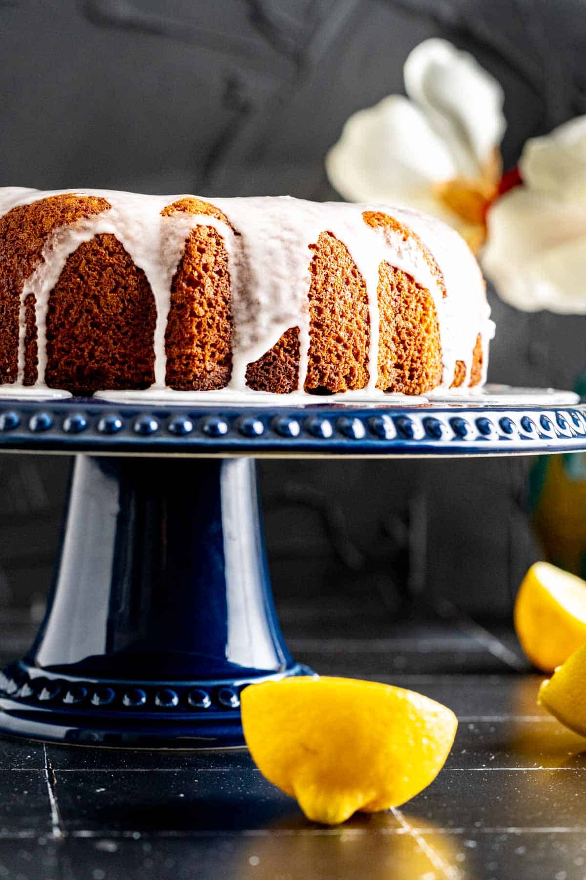 close shot of lemon yogurt cake placed on a cake plate on black tiles with lemon pieces scattered on the table.