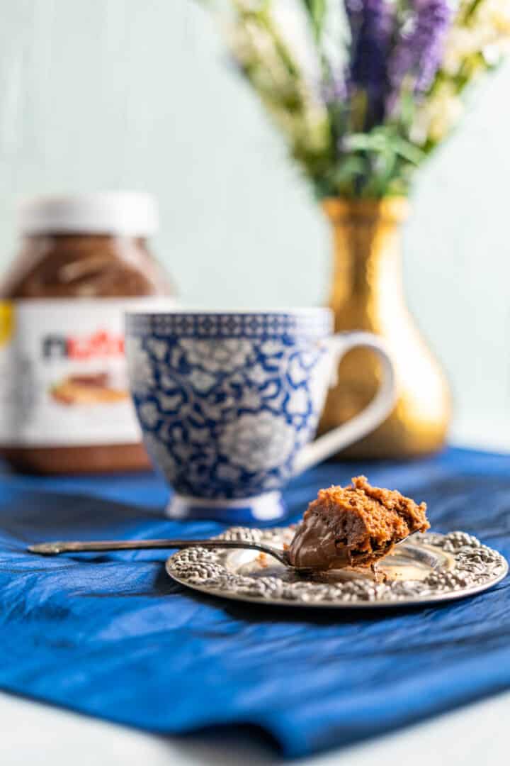 one bite of mug cake in an antique silver plate with flower vase in the backdrop.