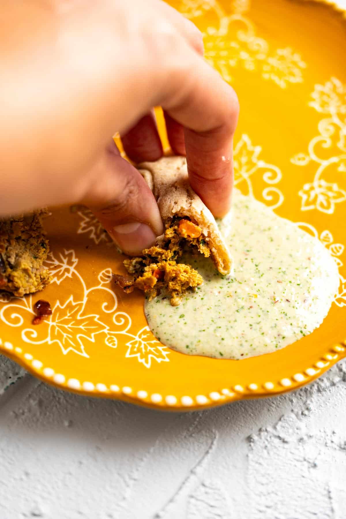 Making a bite of chapli kabab with a pieces of naan and yogurt in a yellow plate.