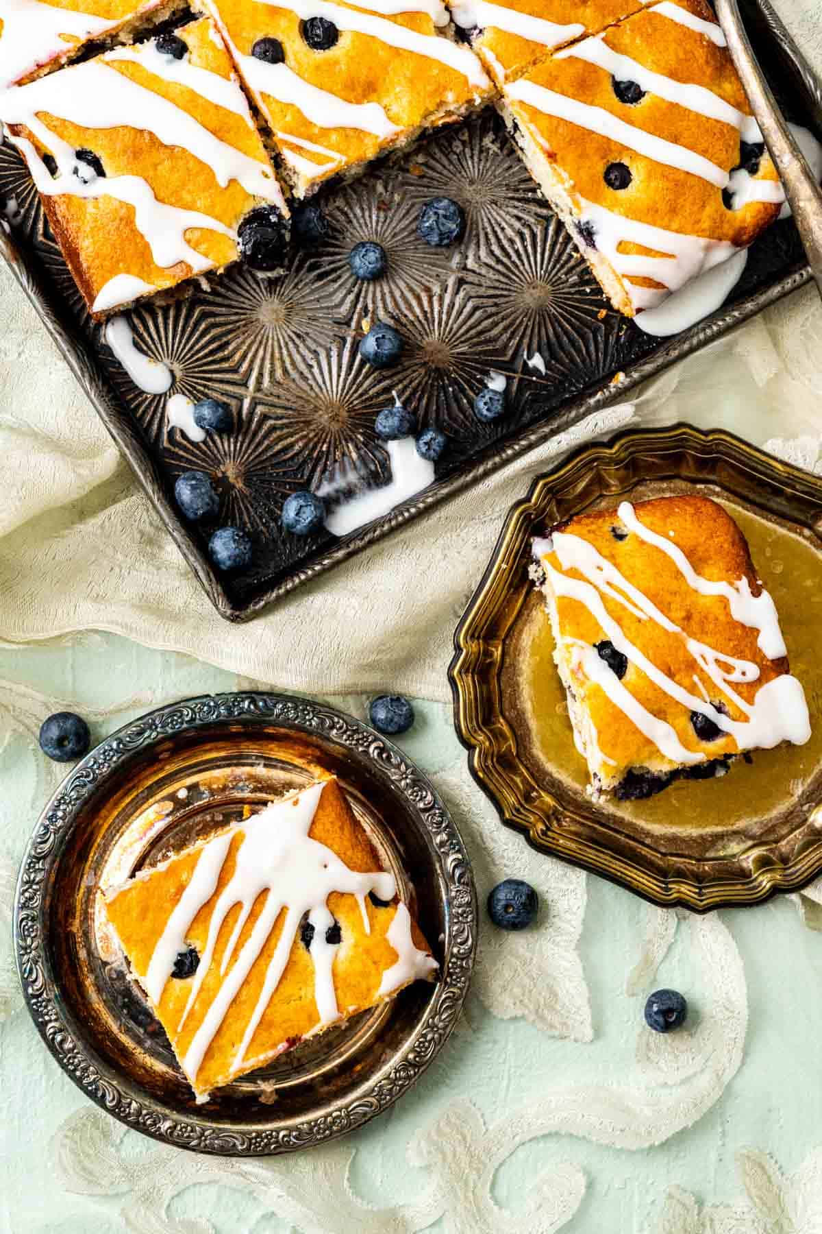 two square pieces of blueberry cake with lemon glaze in two small antique plates with blueberries spread around.