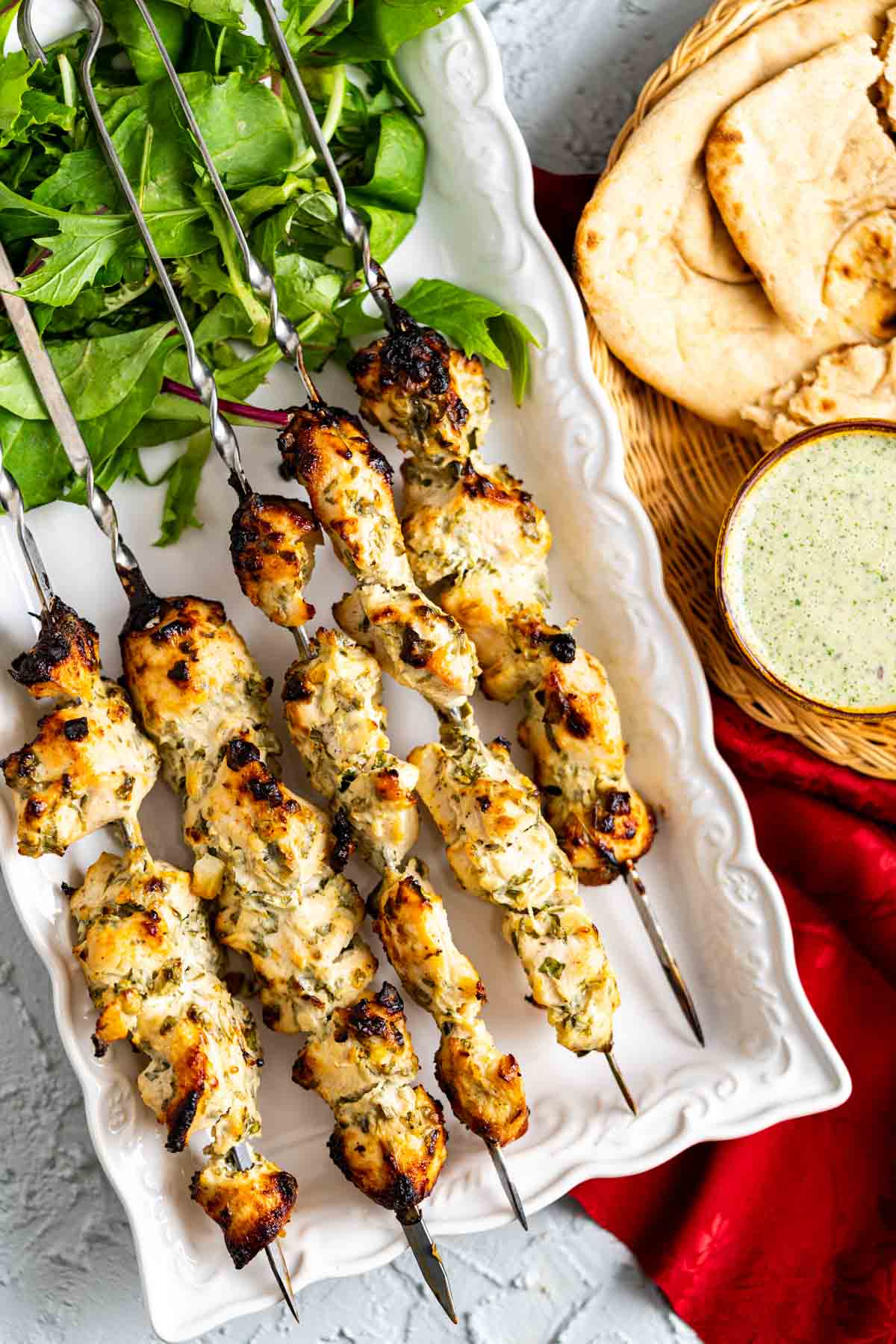 Chicken malai boti skewers in a white plate with salad leaves and mint chutney on the side.