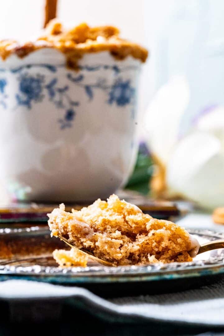 one bite of cinnamon mug cake in an antique silver spoon placed on an antique silver plate with mug in the backdrop.