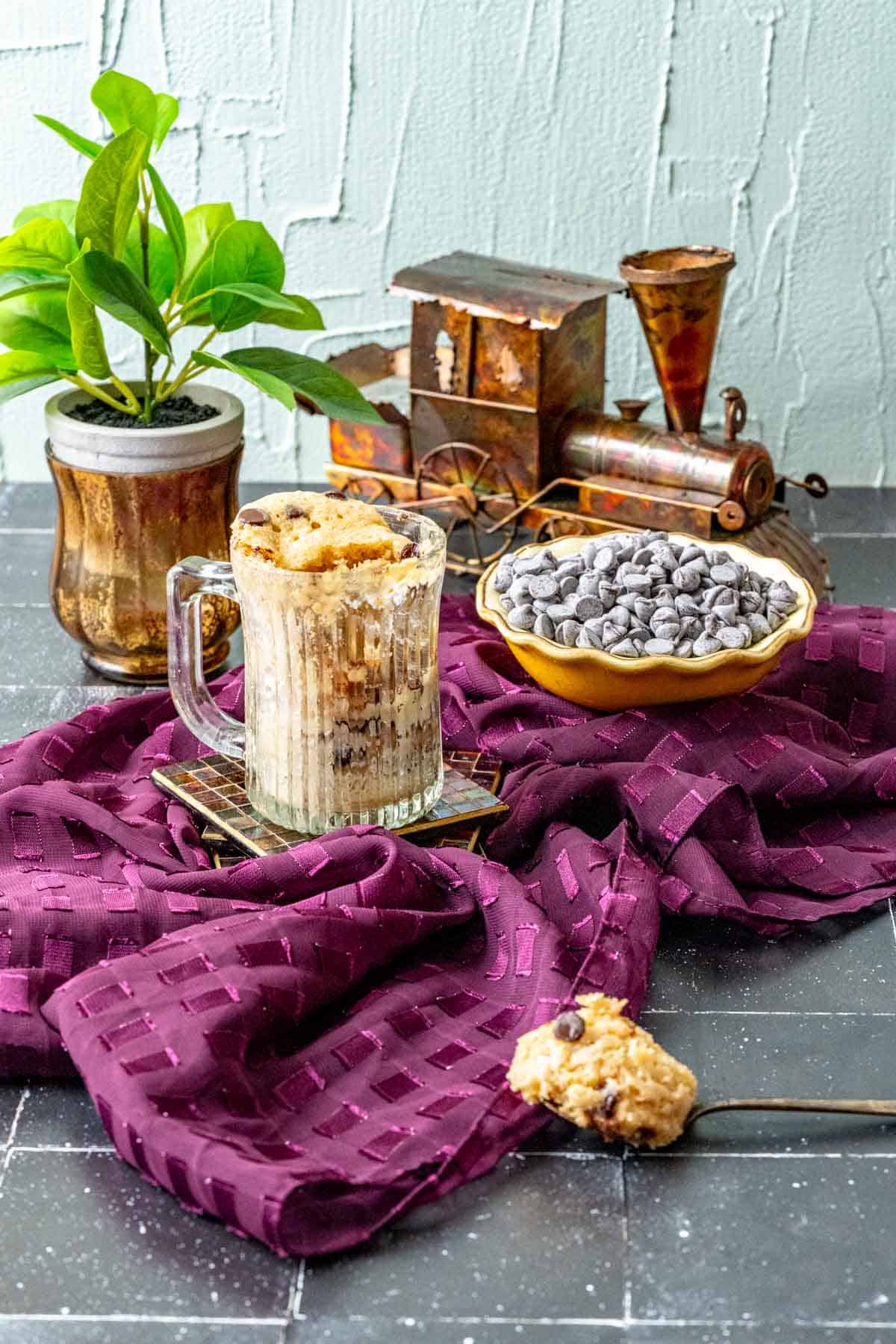 Chocolate chip mug cake sitting on a magenta table cloth with a plant in the backdrop.