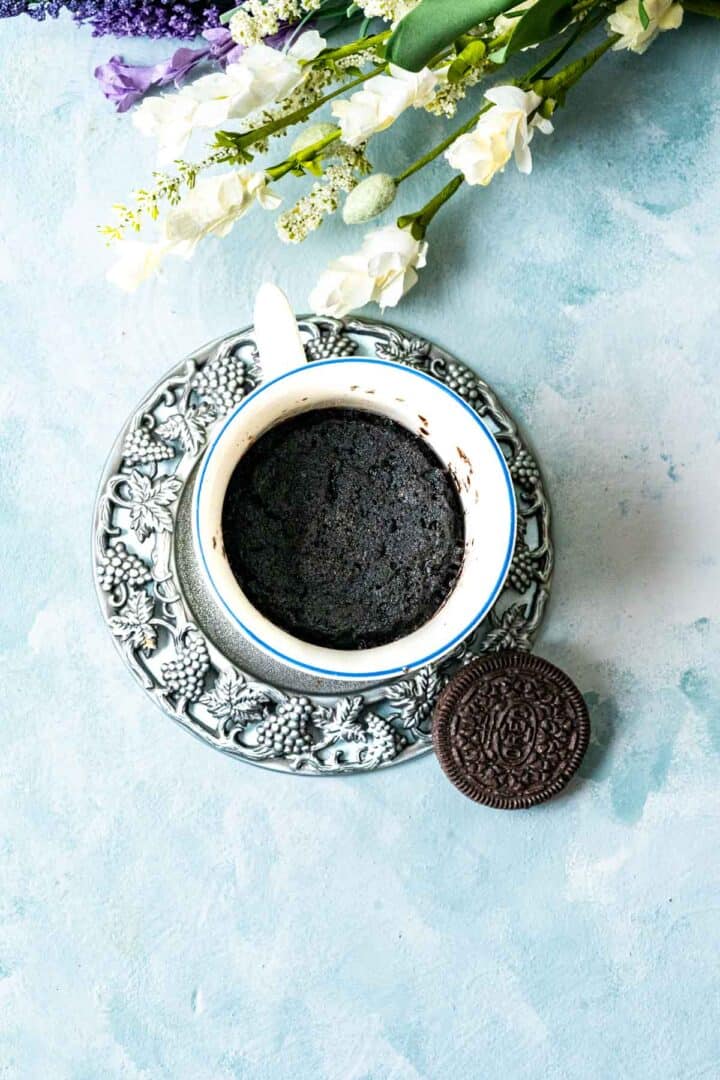 overhead shot of oreo mug cake sitting on blue table and an oreo cookies on the side of a silver plate.