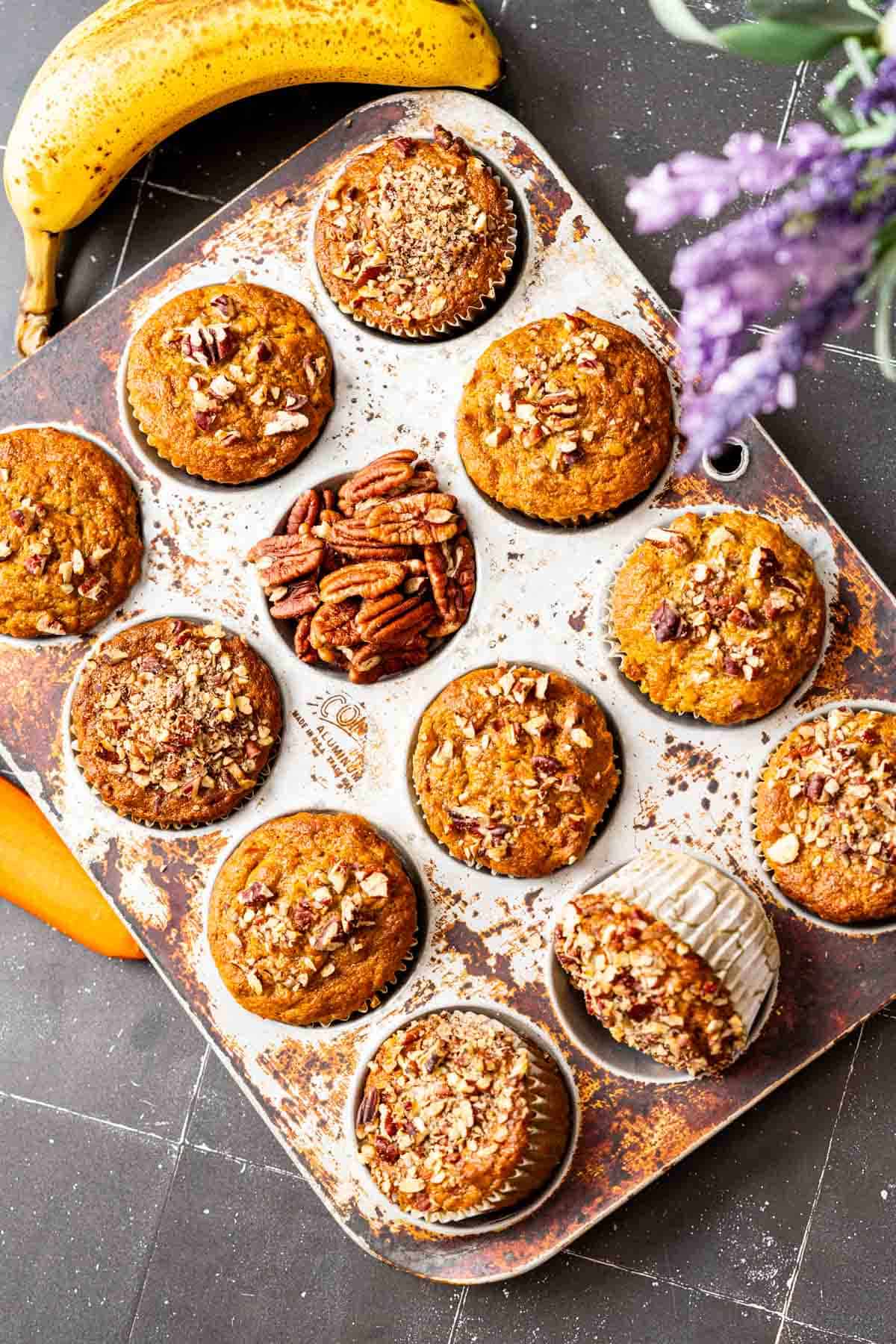 Banana carrot muffins sitting in an antique muffin tray with pecans and banana on the side.