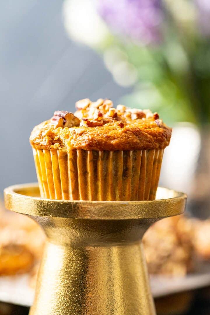 Carrot muffin with banana sitting on a golden stand showing the crust of the muffin with pecans.