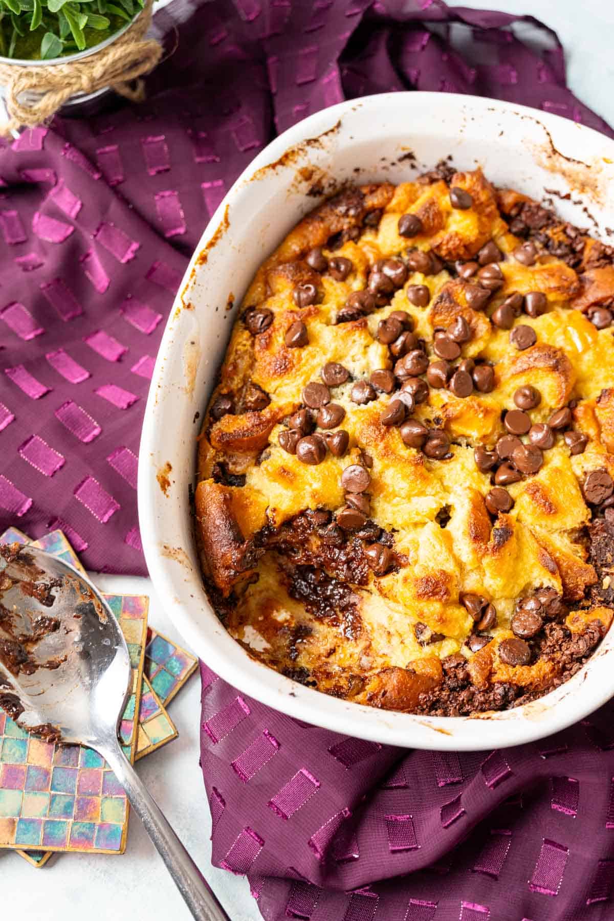 Chocolate chip bread pudding resting on a table with magenta table cloth and a silver serving spoon placed nearby.