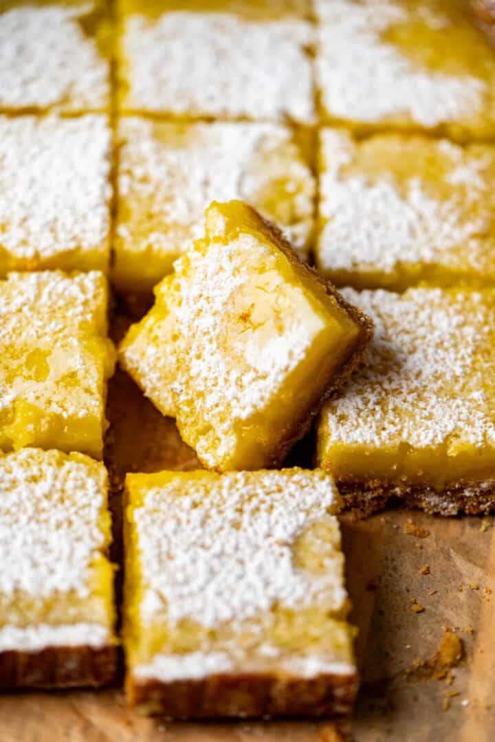 One lemon bar showing the texture of the crust and curd sitting over the rest of the bars on a brown paper.